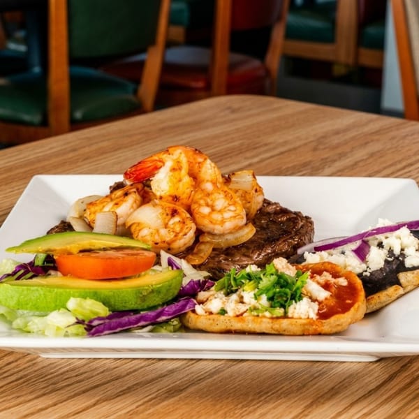 Grilled shrimp and steak with avocado, tomato, and two sopes on a white plate.