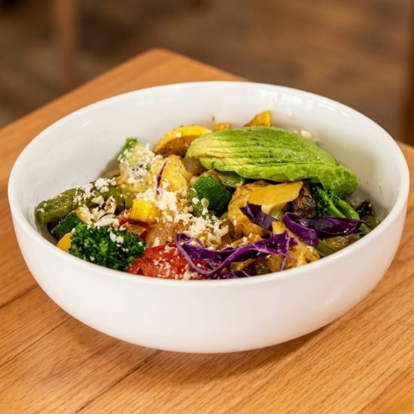 Colorful vegetable bowl with avocado, broccoli, red cabbage, and crumbled cheese on a wooden table.